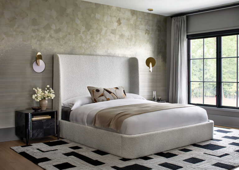 A primary bedroom becomes a serene, quietly sophisticated retreat through a deft combination of layered neutrals and compelling, textural surfaces. A luminous, accent wall in metallic grasscloth is a backdrop for the upholstered platform bed; a geometric accent pillow picks up the pattern in the rug.