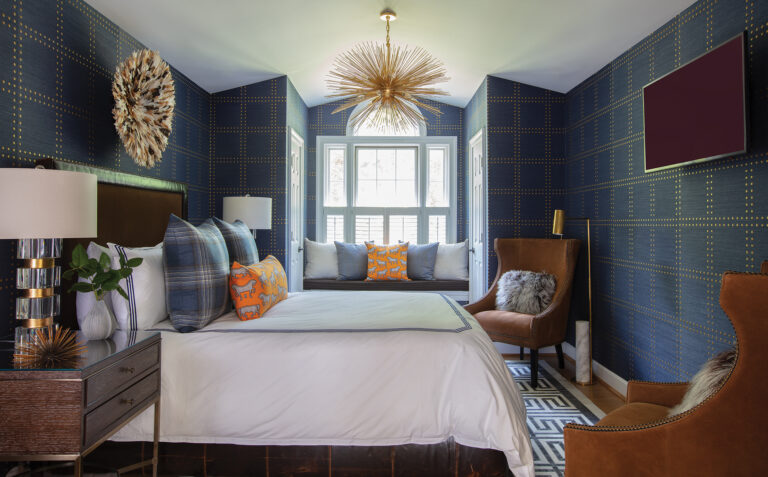 A layered scheme in the bedroom features walls covered in navy grasscloth studded with gold rivets, an eye-catching brass chandelier and orange accents.