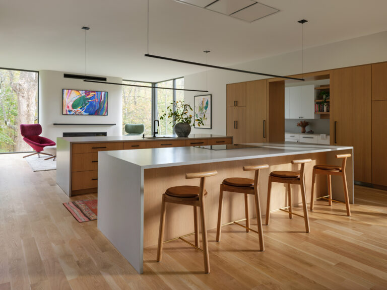 White oak cabinetry, wood and leather bar stools, and a waterfall-edge island unite to command an open kitchen that flows effortlessly into the living area beyond.