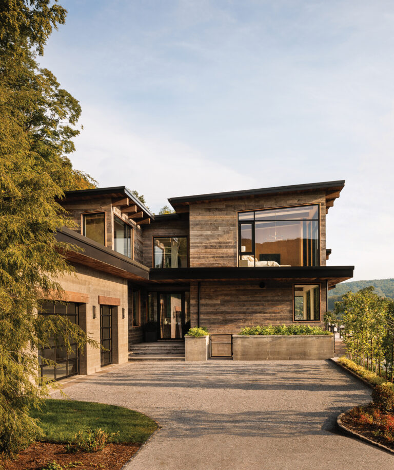 The home’s entry approach reveals board-form concrete and wood cladding, expansive glazing and planted terraces; dramatic Hudson Valley views lie visible beyond.